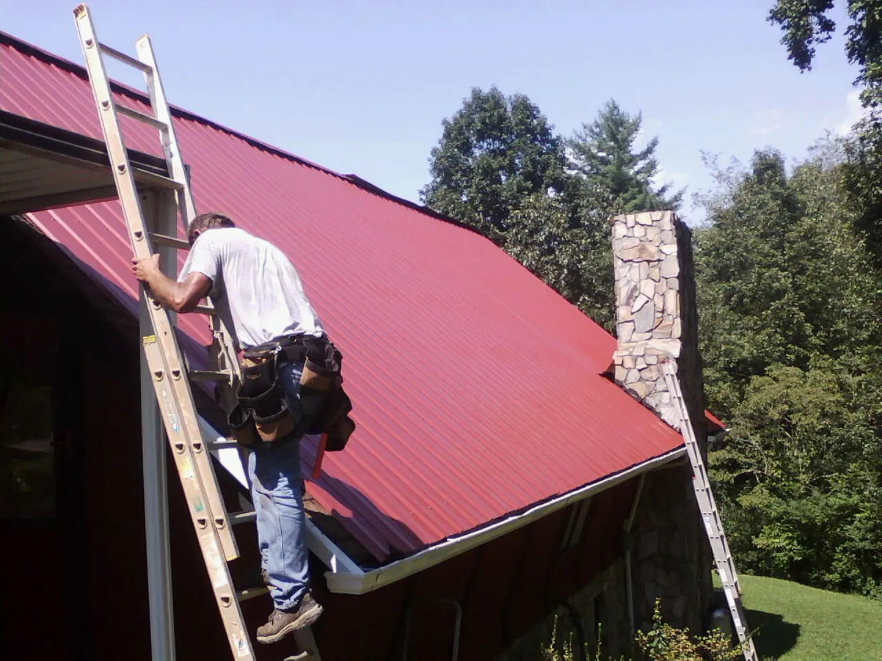 Roofer on a ladder beside a red metal roof and stone chimney — Soffit & Fascia Repair by Roofing Brothers in Cutler
